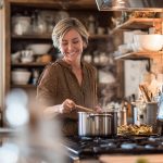 A smiling woman enjoys cooking in her organized, rustic kitchen, stirring a pot on the stove.
