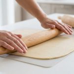 A close-up shows clean hands expertly guiding a smooth, tapered French rolling pin over perfectly uniform, light-colored dough on a minimalist kitchen counter.