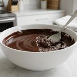 A hyperrealistic close-up shows a white ceramic bowl of melted dark chocolate on a clean kitchen counter. The chocolate is dramatically split: one half is perfectly smooth, glossy, and flowing; the other is thick, grainy, and seized, with a stainless steel spoon stuck in it. A small ramekin of clear liquid sits nearby, hinting at a rescue.