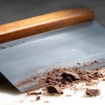 A stainless steel bench scraper with a wood handle chopping dark chocolate on a marble countertop.