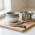 A highly photorealistic still life photograph of a clean, minimalist collection of essential kitchen tools. It features a gleaming stainless steel chef's knife, a light wood cutting board, nesting mixing bowls, a silicone spatula with a light wood handle, a sturdy whisk, and a small modern saucepan. These tools are meticulously arranged on a pristine, light-colored surface, bathed in soft, natural light, creating a fresh, calm, and inviting feel with a neutral color palette.