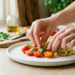 A close-up of gentle hands meticulously arranging vibrant green basil leaves and red cherry tomatoes on a pristine white plate in a softly lit, modern kitchen, conveying quiet culinary artistry.