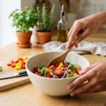 A medium close-up of diverse hands stirring vibrant chopped vegetables in a matte ceramic bowl with a wooden spoon. On a clean, light kitchen counter, a cutting board with more vegetables and a knife are visible, all bathed in soft natural light.