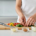 Close-up of human hands meticulously arranging fresh, vibrant ingredients on a clean kitchen countertop. Neatly chopped bell peppers, cherry tomatoes, and herbs are on a cutting board beside a resting chef's knife. Small prep bowls hold pre-portioned ingredients, clearly demonstrating 'mise en place' in a bright, photorealistic, and organized modern kitchen setting.