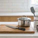 An ultra-photorealistic, minimalist scene of essential cooking tools neatly arranged on a pristine, light kitchen counter. A sharp chef's knife, wooden cutting board, stainless steel saucepan, and measuring cups are prominent, bathed in soft natural light.