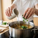A close-up shows a novice cook's hands adding vibrant green parsley and water from a ceramic jug to steaming, golden-brown vegetable soup in a stainless steel pot. The soup's deep color subtly indicates a past culinary error, and the bright, warmly lit kitchen suggests a moment of learning and problem-solving.