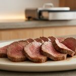 A photorealistic close-up of perfectly cooked, thinly sliced medium-rare roast meat, such as beef tenderloin or pork loin, glistening with natural juices. The elegant slices are artfully arranged on a minimalist ceramic platter, set against a soft-focused modern kitchen background with warm ambient lighting, conveying refined culinary precision and quiet elegance.