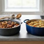 A photorealistic still life featuring an earthy-toned matte stoneware casserole and a gleaming colored enameled cast iron Dutch oven, both filled with impeccably cooked, steaming gourmet food. They rest on a minimalist wood or stone surface, bathed in soft natural light against a softly blurred background.
