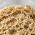 Ultra-photorealistic macro cross-section of a perfectly structured, moist, golden gluten-free crumb. It reveals an intricate, elastic, airy internal matrix where a translucent, glistening, web-like network encases tiny spherical particles and microscopic moisture droplets. Subtle, glowing, holographic molecular models are visible in the soft-focus background, creating a clean, scientific, and appetizing aesthetic.