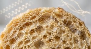 Ultra-photorealistic macro cross-section of a perfectly structured, moist, golden gluten-free crumb. It reveals an intricate, elastic, airy internal matrix where a translucent, glistening, web-like network encases tiny spherical particles and microscopic moisture droplets. Subtle, glowing, holographic molecular models are visible in the soft-focus background, creating a clean, scientific, and appetizing aesthetic.