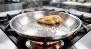 A close-up of a gleaming, superheated stainless steel skillet on a gas stovetop. Perfectly spherical water droplets dance and skitter across the shimmering surface, floating on an invisible cushion of vapor, demonstrating the Leidenfrost effect. Wisps of steam rise from the pan, which also holds a perfectly seared, golden-brown lean protein appearing unbonded and ready to slide freely, all within a clean, professional kitchen aesthetic.