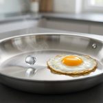 A pristine, highly polished stainless steel skillet on a dark gray counter. A single water droplet hovers on a vapor layer with a wisp of steam on the left. To its right, a perfectly cooked fried egg with a vibrant yolk rests effortlessly, non-stick, on the hot pan. The skillet is reflective with a subtle heat haze against a softly blurred kitchen.