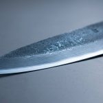 A hyperrealistic macro photograph of a pristine Japanese chef's knife blade with a mirror-polished edge and a deep blue-grey surface, revealing subtle, glowing internal patterns symbolizing its metallurgical properties. The blade rests on a sleek, dark laboratory surface, conveying a clean, scientific aesthetic.