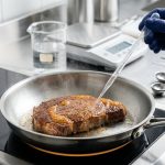A perfectly seared, golden-brown piece of protein sizzles in a pristine stainless steel pan, displaying a uniform, crisp golden-amber crust. A gloved hand delicately mists the surface with a clear liquid from a sleek spray bottle, enhancing its sheen. In the softly blurred background, organized scientific culinary tools like a beaker, digital scale, and measuring spoons are visible. Bright, analytical lighting illuminates the modern kitchen/lab setting, with subtle steam rising and a warm glow beneath the pan, conveying scientific precision.