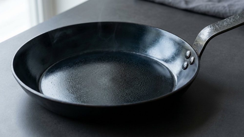 A hyper-photorealistic close-up of a perfectly seasoned, dark carbon steel skillet. Its interior surface has a deep, lustrous, near-mirror-like sheen, reflecting light like a well-maintained nonstick surface. A faint, shimmering heat distortion is visible just above the rim, set against a clean, minimalist background.