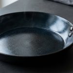 A hyper-photorealistic close-up of a perfectly seasoned, dark carbon steel skillet. Its interior surface has a deep, lustrous, near-mirror-like sheen, reflecting light like a well-maintained nonstick surface. A faint, shimmering heat distortion is visible just above the rim, set against a clean, minimalist background.