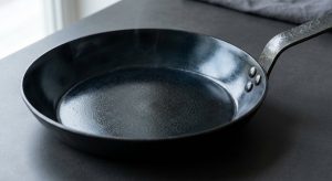 A hyper-photorealistic close-up of a perfectly seasoned, dark carbon steel skillet. Its interior surface has a deep, lustrous, near-mirror-like sheen, reflecting light like a well-maintained nonstick surface. A faint, shimmering heat distortion is visible just above the rim, set against a clean, minimalist background.