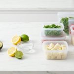 A clean, hyper-realistic still life photograph on a light kitchen counter, contrasting initial enzymatic browning with effective preservation. On one side, green apple slices and a halved avocado show faint discoloration, alongside fresh lemon and lime halves and an acidic solution. On the other, vibrant, perfectly fresh chopped basil, bell pepper strips, potato cubes, and a fish fillet are impeccably preserved in transparent silicone containers.