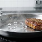 A hyper-realistic close-up of a hot stainless steel skillet, showing spherical water droplets hovering and dancing due to the Leidenfrost effect, next to a perfectly seared golden-brown protein with a crisp crust. A subtle heat haze rises from the pan.