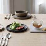 A photorealistic, close-up view of a sophisticated, minimalist dining table setting. Exquisitely plated individual gourmet dishes showcase vibrant acidic elements next to rich savory components, and crisp cool vegetables paired with warm proteins. They are served on a curated selection of high-grade vessels, including matte ceramic plates, polished stainless steel cutlery, clear borosilicate glass, and smooth slate or light wood boards. Soft, natural diffused light highlights fine textures and subtle reflections against a softly blurred elegant background, conveying refined culinary artistry.