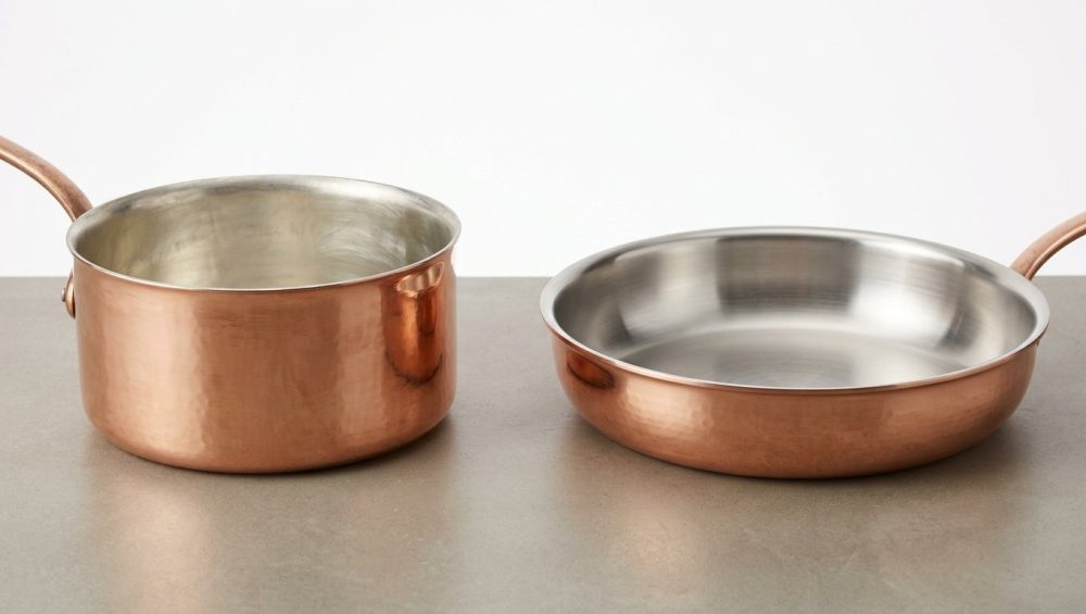 A photorealistic studio shot showcases two exquisite hand-forged copper pans on a clean, neutral surface. A deep saucepan features a soft, matte tin lining, while a shallow sauté pan displays a bright, reflective stainless steel lining. The image highlights the distinct textures and finishes of both interiors against the warm, hammered copper exteriors.