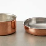 A photorealistic studio shot showcases two exquisite hand-forged copper pans on a clean, neutral surface. A deep saucepan features a soft, matte tin lining, while a shallow sauté pan displays a bright, reflective stainless steel lining. The image highlights the distinct textures and finishes of both interiors against the warm, hammered copper exteriors.