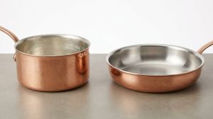 A photorealistic studio shot showcases two exquisite hand-forged copper pans on a clean, neutral surface. A deep saucepan features a soft, matte tin lining, while a shallow sauté pan displays a bright, reflective stainless steel lining. The image highlights the distinct textures and finishes of both interiors against the warm, hammered copper exteriors.
