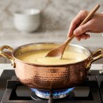 A hyper-detailed close-up of a pristine, hand-hammered copper saucepan, gleaming under soft light, as a chef's hand gently stirs a perfectly smooth, creamy pale yellow sauce inside with a wooden spoon, over a low blue gas flame in a high-end kitchen, conveying culinary precision and luxury.