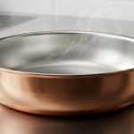 A photorealistic close-up, looking down into a gleaming, polished copper pan with a pristine, silvery tin interior. A subtle heat shimmer rises from the perfectly smooth tin. The pan sits on a dark, minimalist surface, bathed in soft studio light, highlighting its unblemished quality and scientific elegance.