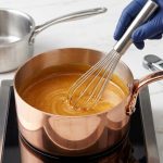A hyper-realistic close-up of a gleaming copper saucepan filled with shimmering golden caramel being stirred by a whisk. A digital thermometer and sugar crystals rest on a white marble counter, with a blurred stainless steel pot in the background, conveying culinary precision.