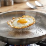 A photorealistic close-up of a perfectly cooked fried egg effortlessly sliding across a pristine, uncoated stainless steel frying pan. A visible steam cushion prevents adhesion beneath the egg, while nearby dancing water droplets demonstrate the Leidenfrost effect on the intensely hot metal.