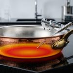 A photorealistic, close-up image of an exquisite, hand-hammered copper sauté pan centered on a sleek, dark reflective laboratory surface. Its polished copper exterior, with a classic brass handle, shows subtle hammered textures. A subtle thermal imaging overlay of reds and yellows depicts perfectly even heat distribution across its base. The pan's interior reveals a pristine, silvery tin lining, while precision metallurgical instruments, like a digital caliper, rest on its rim. The scene is rendered with razor-sharp detail and dramatic, high-contrast lighting, set against a softly blurred, minimalist high-tech lab background, conveying scientific rigor and premium craftsmanship.