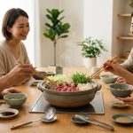 An ultra-photorealistic, high-detail image of a serene communal dining experience. At the center, a gently steaming Japanese donabe pot, filled with Wagyu, fresh vegetables, and udon, rests on a modern wood table with an integrated cooktop. Around the table, three to four adults mindfully use chopsticks, smiling and conversing in soft, natural daylight, conveying a sense of shared well-being.