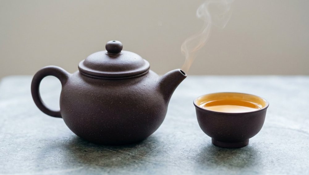 A hyper-realistic studio shot of an unglazed, purplish-brown Yixing clay teapot with a wisp of steam rising from its spout, and a matching teacup partially filled with amber-golden tea. The textured teaware rests on a light neutral surface against a clean, softly diffused background.