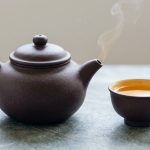 A hyper-realistic studio shot of an unglazed, purplish-brown Yixing clay teapot with a wisp of steam rising from its spout, and a matching teacup partially filled with amber-golden tea. The textured teaware rests on a light neutral surface against a clean, softly diffused background.