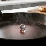 A perfectly spherical water droplet levitates and dances on an intensely hot, dark frying pan with wisps of steam, demonstrating the Leidenfrost effect. In the soft-focus background, a golden-brown seared piece of meat rests on a wooden cutting board.