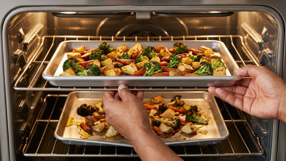 Close-up: An adult's hands slide an impeccably flat sheet pan, filled with perfectly roasted broccoli, golden potatoes, and caramelized carrots, into a modern oven. On a lower, blurry rack, a severely warped pan with unevenly cooked, oily vegetables provides a stark visual contrast.