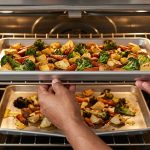 Close-up: An adult's hands slide an impeccably flat sheet pan, filled with perfectly roasted broccoli, golden potatoes, and caramelized carrots, into a modern oven. On a lower, blurry rack, a severely warped pan with unevenly cooked, oily vegetables provides a stark visual contrast.