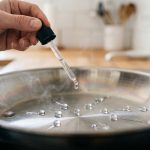 A photorealistic close-up of a pristine stainless steel skillet, perfectly heated to demonstrate the Leidenfrost effect. Spherical water droplets skitter and dance across the hot surface, surrounded by delicate steam, as a skilled hand carefully releases a final water drop. A softly blurred, modern kitchen forms the background, emphasizing precision and scientific elegance.