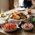 A photorealistic, elegantly arranged build-your-own crostini station, bathed in soft natural light. Golden-brown, crisp crostini are stacked in a rustic basket, surrounded by small bowls of vibrant toppings including diced tomatoes, creamy ricotta, prosciutto, fresh basil, Kalamata olives, and roasted red pepper spread. The detailed food is in sharp focus against a softly blurred background.