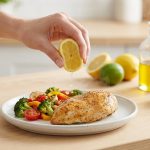 A close-up of a hand gracefully squeezing a vibrant half-lemon over a warm, roasted chicken breast, with glistening juice droplets arcing towards the food. In the soft-focus background, a pristine kitchen counter holds more fresh citrus and bottles, all bathed in bright natural light.