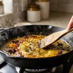 A close-up, photorealistic shot of vibrant dry spices actively blooming and sizzling in hot golden oil in a dark frying pan. A blurred hand gently stirs with a wooden spoon as aromatic steam rises in a warm, inviting kitchen setting.