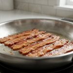 A close-up shows perfectly cooked, golden-brown crispy bacon with glistening rendered fat in a stainless steel or cast iron frying pan, on a clean kitchen stovetop under soft natural light.