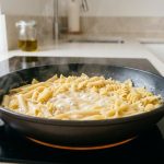 Close-up of mixed dried pasta simmering in a shallow non-stick skillet on a modern induction stovetop. A thick, glossy starchy liquid surrounds the pasta, with gentle steam rising. The background features a bright, minimalist kitchen countertop.