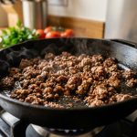 A photorealistic close-up shows perfectly browned ground beef searing in a heavy cast iron skillet. Distinct, crispy caramel-brown pieces of meat emit subtle wisps of steam, set against a softly blurred modern kitchen background with hints of fresh ingredients.