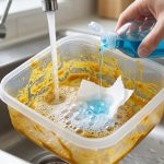 A clean hand pours blue dish soap into a clear plastic food container stained with yellow-orange oil residue. The container sits in a modern stainless steel sink with warm water and a torn paper towel, showing the start of a cleaning process.