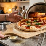 A casual home cook confidently slides a vibrant, artisanal homemade pizza, dusted with semolina flour, off a smooth wooden peel and into the glowing opening of a modern home pizza oven. The perfectly shaped dough showcases bright red sauce, creamy melted mozzarella, and fragrant green basil, all illuminated by warm, natural light.