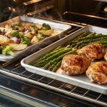Inside a modern oven, two baking sheets illustrate a comparison: a thin, warped pan holds unevenly roasted vegetables with burnt and pale spots, while a thick, heavy-gauge pan displays perfectly and evenly cooked gourmet ingredients.