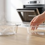 Two glass baking dishes sit side-by-side on a modern kitchen counter. The dish on the left is heavily fractured with a prominent spiderweb crack pattern and subtle wisps of steam, suggesting thermal shock. In sharp contrast, the borosilicate glass dish on the right is pristine and intact. A pair of hands gently reaches towards the undamaged borosilicate dish, with a stainless steel oven slightly ajar in the soft-focus background.