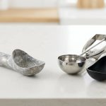 A photorealistic image on a clean kitchen counter showcases three ice cream scoops, contrasting damaged and durable tools. On the left, a dull, corroded aluminum scoop is visibly mottled with chalky gray residue. To the right, two pristine alternatives gleam: one polished stainless steel, the other a sleek black non-stick design.